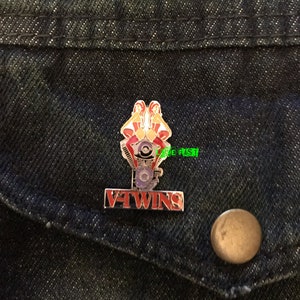 May include: A red and gold pin with two women sitting on a motorcycle engine. The pin says "V-Twins" in red letters.