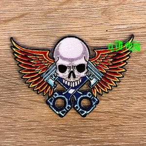 May include: A white skull with red and yellow wings and two crossed blue pistons. The skull has a black eye socket and a toothy grin.