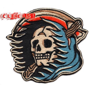 May include: Embroidered patch featuring a skull with a blue and white bandana, surrounded by flames. The skull has a white face and black eyes. The text "Live Fast" is visible in the top left corner.