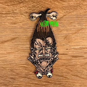 May include: Embroidered patch depicting a cartoon character with a bone-shaped hairstyle, a large moustache, and a menacing expression. The character is wearing a red and white outfit and has a bone-like accessory on each hand.