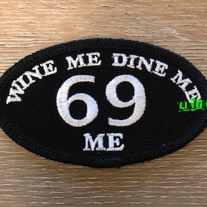 May include: Black oval patch with white embroidered text that reads "Wine Me Dine Me 69 Me".