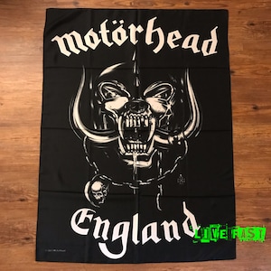 Pode incluir: Bandeira retangular preta com o nome da banda "Motörhead" em letras góticas brancas acima de uma ilustração detalhada de uma caveira com presas e correntes. A palavra "England" está abaixo da caveira. A frase "LIVE FAST" está em verde.