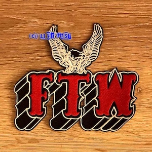 Op de afbeelding: Een geborduurde patch met de letters "FTW" in een vet, gestileerd lettertype. Een witte adelaar met uitgespreide vleugels bevindt zich boven de letters. De patch is rood, wit en zwart.