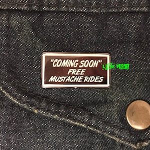 May include: A silver enamel pin with a white border and black background. The text on the pin reads "Coming Soon" Free Mustache Rides.