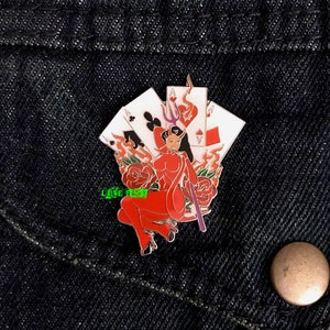 May include: A red and black enamel pin featuring a devil woman holding a trident and surrounded by playing cards and roses. The pin has a shiny finish and is perfect for adding a touch of whimsy to any outfit.