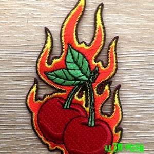 May include: Embroidered patch featuring two red cherries on fire. The cherries have green stems and leaves. The flames are orange and yellow.
