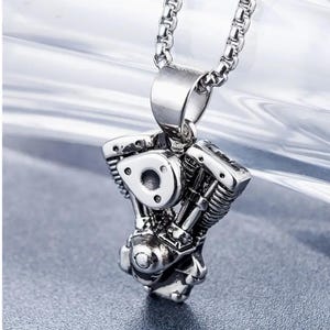 Puede incluir: Un collar con un colgante de motor de motocicleta plateado. El colgante es detallado y realista, con pistones y cilindros visibles. La cadena es una cadena de caja plateada.