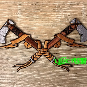 May include: Two crossed tomahawks with brown handles and white blades. The handles are wrapped in a braided design and have a brown and orange fringe.