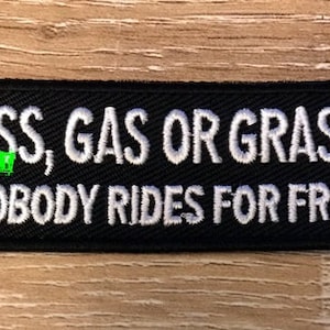 Puede incluir: Parche rectangular negro con texto bordado en blanco: "ASS, GAS OR GRASS NOBODY RIDES FOR FREE". El parche tiene un borde negro y está sobre un fondo de vetas de madera.