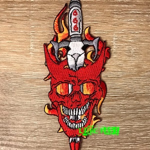 Könnte beinhalten: Roter gestickter Patch mit einem Totenkopf und einem Dolch. Aus dem Schädel schlagen Flammen und der Dolch trägt die Zahl 666.