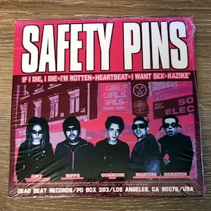 May include: A pink and white album cover for the band Safety Pins. The album cover features a band photo of five people with the band's name in large white letters. The album title is "If I Die, I Die I'm Rotten Heartbeat I Want Sex Kazike".