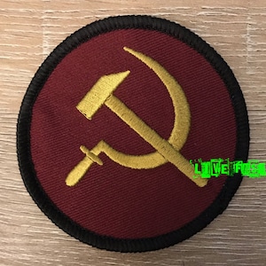 May include: A red embroidered patch with a gold hammer and sickle design. The patch is circular and has a black border.