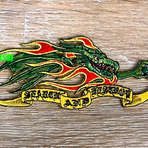 May include: Embroidered patch featuring a green dragon with red and yellow flames. The dragon holds a silver object in its claw. A yellow banner below reads "SEARCH AND DESTROY" in black lettering. The patch is on a wood grain background.