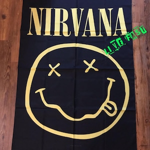 May include: Black fabric banner featuring the word "NIRVANA" in yellow capital letters above a yellow smiley face with crossed-out eyes and a protruding tongue. The banner is rectangular and made of fabric.