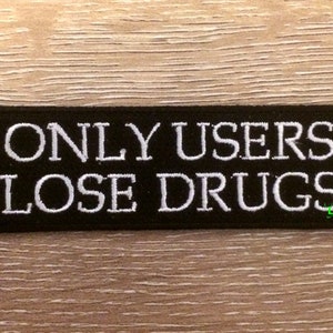 Puede incluir: Parche rectangular negro con texto bordado blanco: "ONLY USERS LOSE DRUGS". El parche está diseñado para ser cosido o planchado en ropa o accesorios. El fondo es una superficie de madera marrón claro.