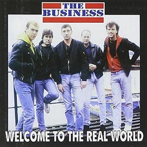 以下が含まれることがあります： 「ザ・ビジネス」というバンドの5人の男性がレンガの壁の前に立っています。バンドメンバーはジーンズとジャケットを着ています。バンド名は、男性の上の看板に赤、白、青の文字で書かれています。画像の下部には、黒い背景に白い文字で「Welcome to the Real World」と書かれています。