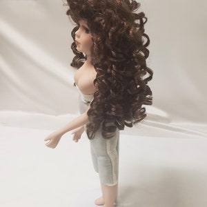 May include: A porcelain doll with brown curly hair and a white dress. The doll is standing on a white stand.
