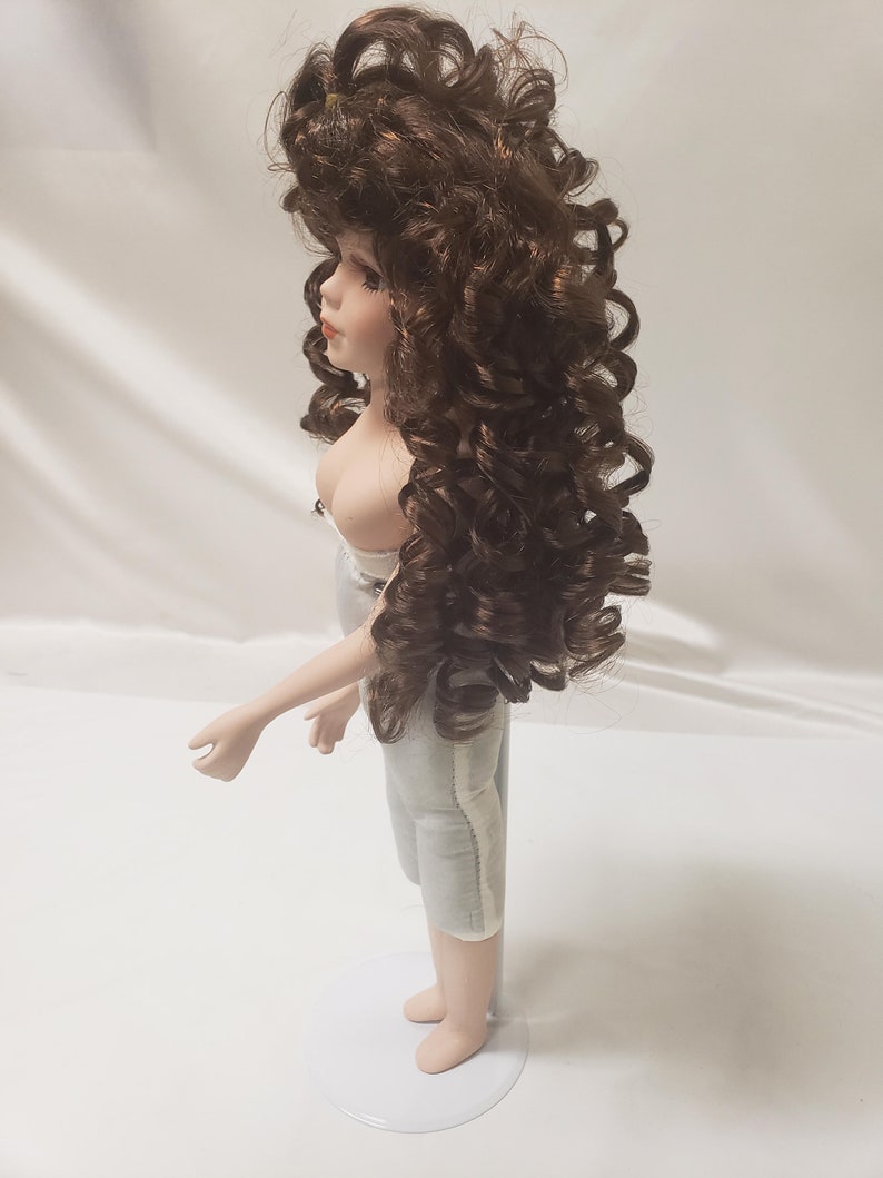 May include: A porcelain doll with brown curly hair and a white dress. The doll is standing on a white stand.