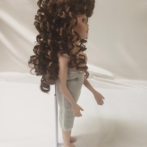 May include: A porcelain doll with brown curly hair, wearing a white dress, standing on a white stand.