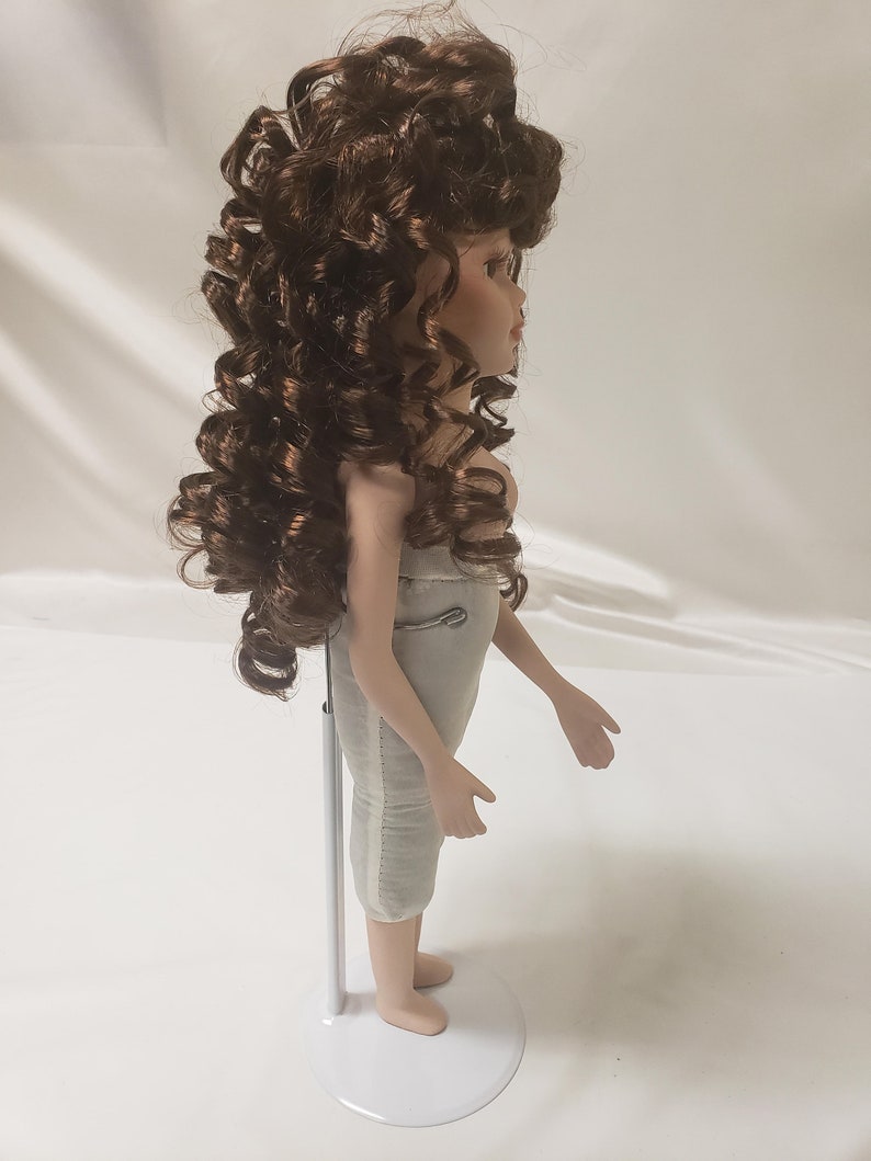 May include: A porcelain doll with brown curly hair, wearing a white dress, standing on a white stand.
