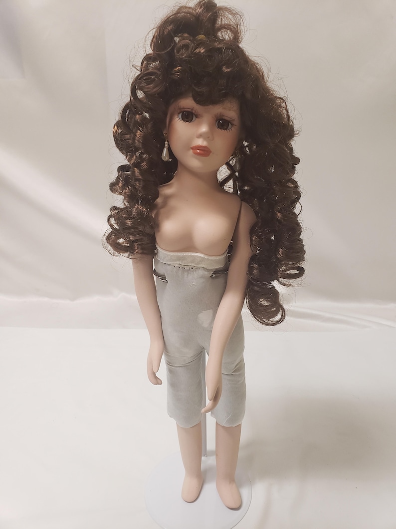 May include: A porcelain doll with brown curly hair, wearing a light gray outfit and standing on a white stand.