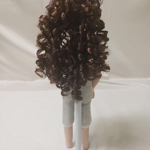May include: A porcelain doll with long, curly brown hair, wearing a light gray outfit and standing on a white stand.