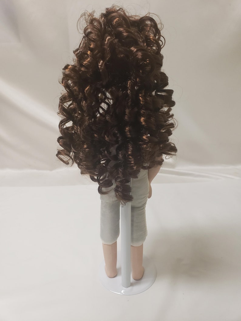 May include: A porcelain doll with long, curly brown hair, wearing a light gray outfit and standing on a white stand.