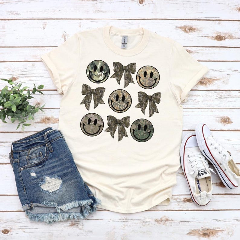 May include: A cream-colored t-shirt with a pattern of camouflage smiley faces and bows. The smiley faces and bows are in shades of green and brown.