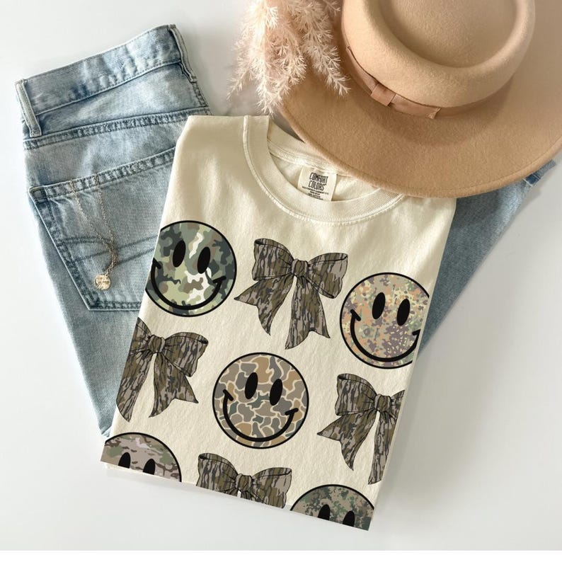 May include: A cream-colored t-shirt with a camouflage print of smiley faces and bows. The shirt has a crew neck and short sleeves.