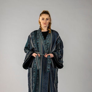 Puede incluir: Una bata larga de color azul y verde con estampado geométrico y ribete blanco. La bata tiene un cinturón para atar. La modelo lleva pantalones negros y botas de tacón negras.