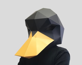 May include: A black and gold papercraft mask shaped like a duck's head. The mask has a geometric design and is worn by a person with their arms crossed.