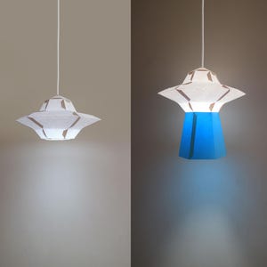 Op de afbeelding: Twee hangende lampen met witte papieren lampenkappen in de vorm van vliegende schotels. De linker lamp is uit en de rechter lamp is aan en verlicht een blauwe papieren lampenkap.