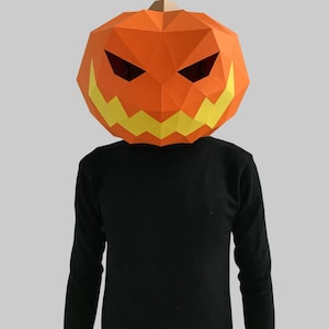Peut inclure: Une personne portant un masque de citrouille en papier m&acirc;ch&eacute; avec un visage effrayant. Le masque est orange avec des dents jaunes et des yeux noirs.
