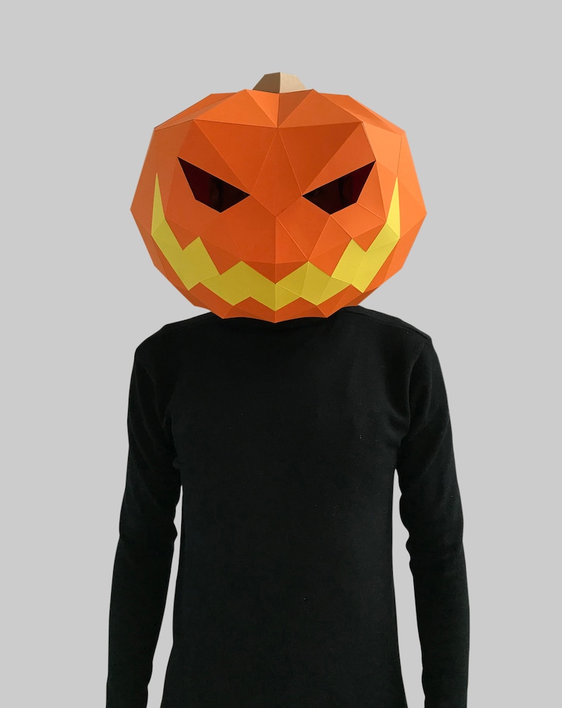 Peut inclure: Une personne portant un masque de citrouille en papier m&acirc;ch&eacute; avec un visage effrayant. Le masque est orange avec des dents jaunes et des yeux noirs.