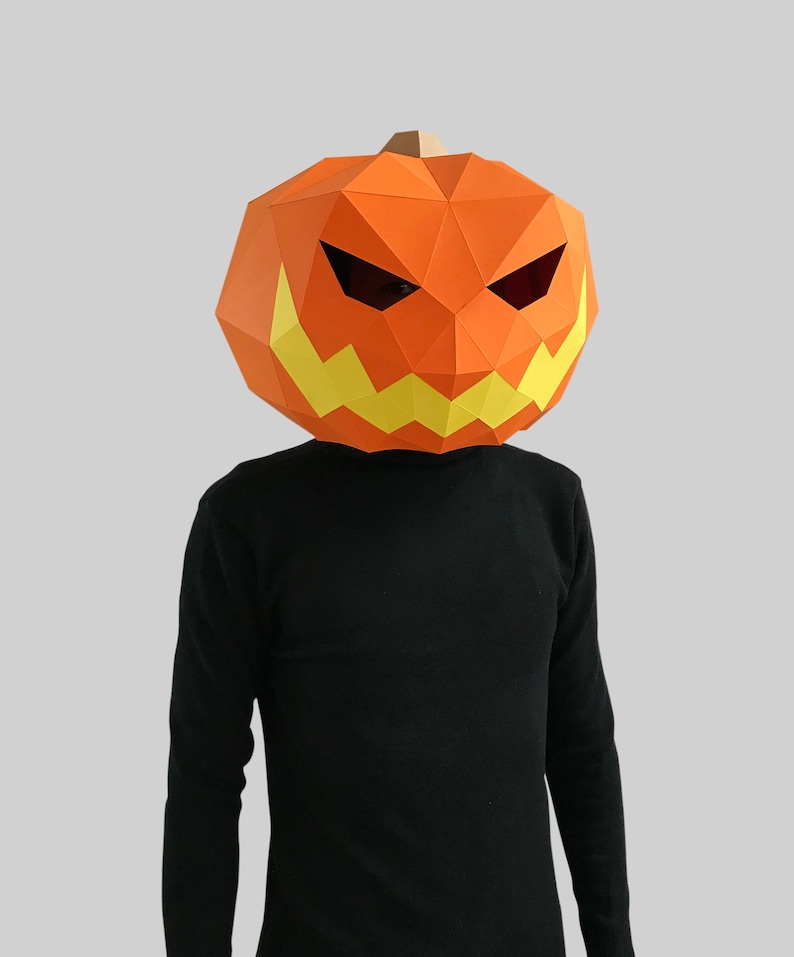 Peut inclure: Une personne portant un masque de citrouille en papier m&acirc;ch&eacute; avec un visage effrayant. Le masque est orange avec des dents jaunes et des yeux noirs.