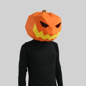 Peut inclure: Une personne portant un masque de citrouille en papier m&acirc;ch&eacute; avec un visage effrayant. Le masque est orange avec des dents jaunes et des yeux noirs.