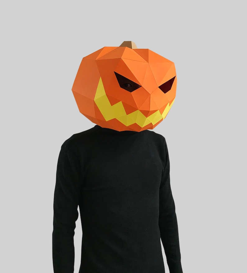 Peut inclure: Une personne portant un masque de citrouille en papier m&acirc;ch&eacute; avec un visage effrayant. Le masque est orange avec des dents jaunes et des yeux noirs.