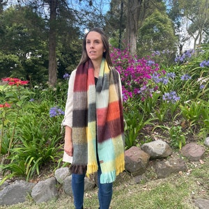 May include: A woman wearing a multi-colored plaid scarf with brown, red, yellow, and green stripes. She is standing in a garden with green grass and flowers.