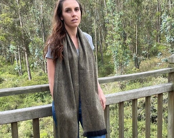 May include: A woman wearing blue jeans and brown ankle boots is standing on a wooden deck. She is wearing a green and black scarf with a fringed edge.