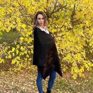 May include: A woman wearing a black shawl and blue jeans stands in front of a tree with yellow leaves. She is wearing black ankle boots.