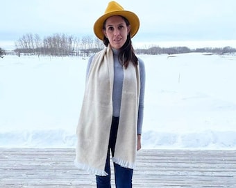 May include: A woman wearing a grey turtleneck sweater, blue jeans, and a white scarf. She is wearing a yellow fedora hat and black ankle boots. She is standing on a wooden deck in front of a snowy field.