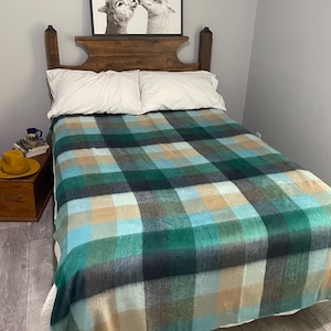 May include: A bed with a wooden headboard, white pillows, and a plaid blanket. The blanket has a pattern of green, blue, and beige squares. A framed black and white picture of two alpacas kissing hangs above the bed.