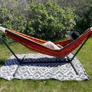 May include: A red, orange, and green striped hammock with fringe hanging from a black metal stand. A person is lying in the hammock wearing a white shirt and a black hat.