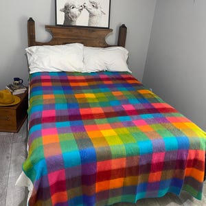 May include: A vibrant, multi-coloured plaid blanket, featuring squares of blue, green, orange, purple, and pink, covers a bed with white pillows. The blanket has a large grid pattern. A wooden headboard and a framed picture of two alpacas are above the bed.