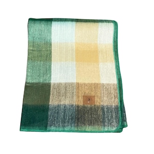 May include: A folded rectangular throw blanket featuring a large plaid pattern in shades of forest green, cream, and soft yellow. The blanket has a finished green stitched edge and a small brown rectangular leather-like patch on the lower right.