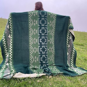 May include: A green and gray patterned blanket with a geometric design. The blanket features a central panel of light green and dark green, with a repeating pattern. The blanket is displayed outdoors on a grassy hill.