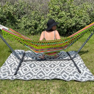 May include: A person relaxing in a colorful striped hammock with fringe, suspended from a black metal stand. The hammock is on a gray and white patterned rug, outdoors on a grassy lawn.