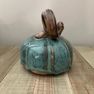 May include: A ceramic pumpkin sculpture featuring a glossy teal glaze with dark blue and brown speckles. The pumpkin has a textured, curved brown stem and visible vertical ridges, with a raw, unglazed tan clay base visible at the bottom.