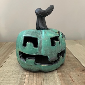 May include: A teal ceramic pumpkin lantern with a black stem and carved facial features including square eyes and a wide mouth. The surface has a distressed finish with dark shading in the crevices and visible black stitching details on the side.