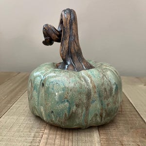 Könnte beinhalten: Eine Kürbisskulptur aus Keramik mit einem runden, gerippten Körper und einer gesprenkelten Glasur in Salbeigrün und Braun. Oben befindet sich ein hoher, strukturierter, dunkelbrauner Stiel, der sich leicht zur Seite neigt und feine Rillen zeigt.
